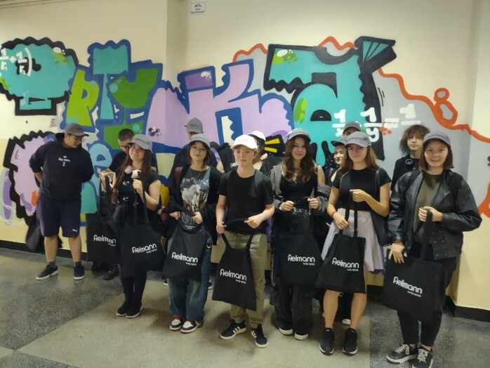 Grupa dzieci stoi na tle ściany z kolorowym grafitii (napis optyka), dzieci trzymają płócienne czarne torby